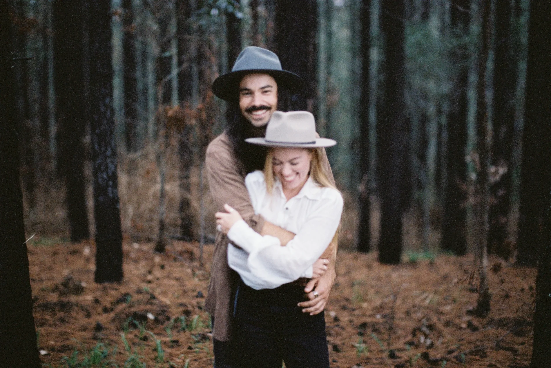 Two people in wide-brimmed hats embracing in a pine forest
