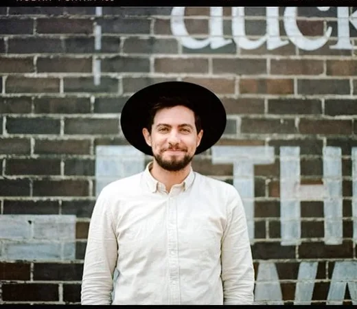 Joshua Mikhaiel in a wide-brimmed hat against a brick wall